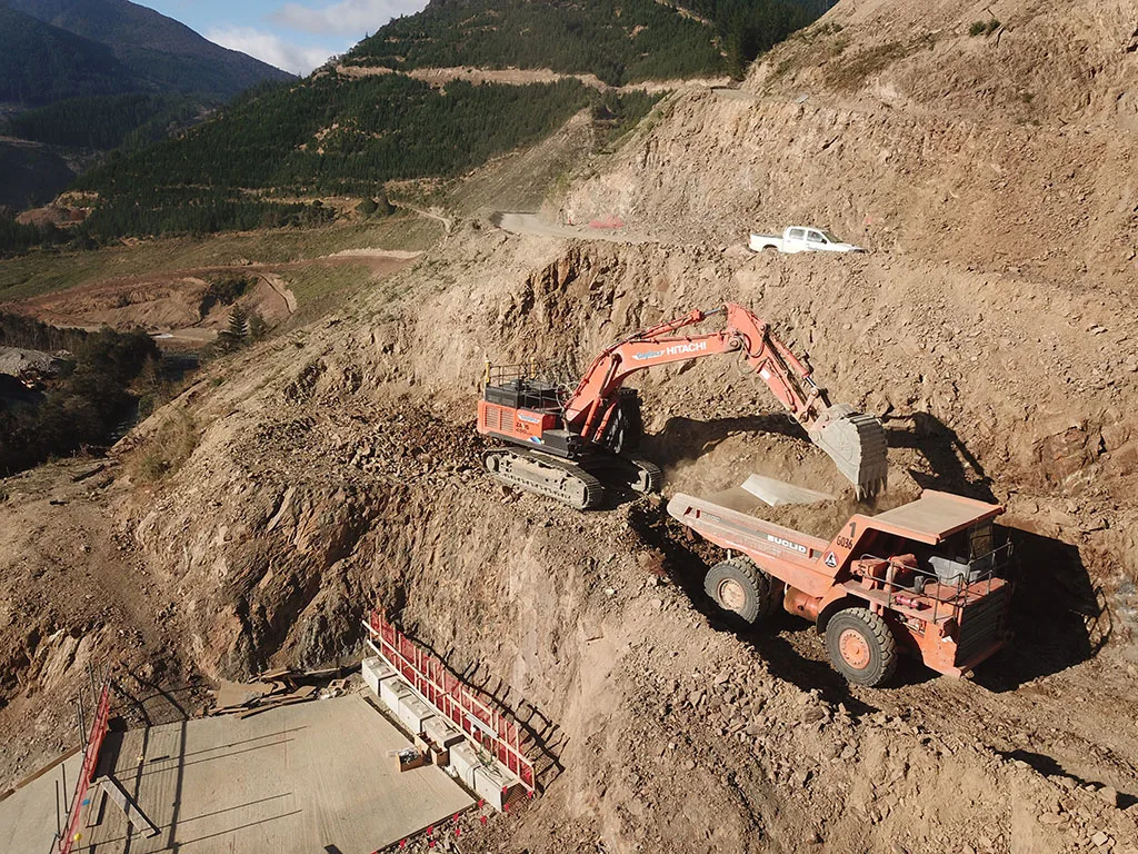 Excavator loading dirt into a large truck at a construction site with a mountain backdrop.