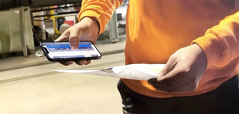 Worker in orange shirt using smartphone and holding paper documents.