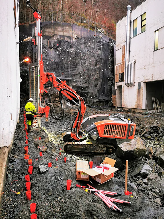 Construction worker operating a drill rig next to a rock face with a mini excavator on site.