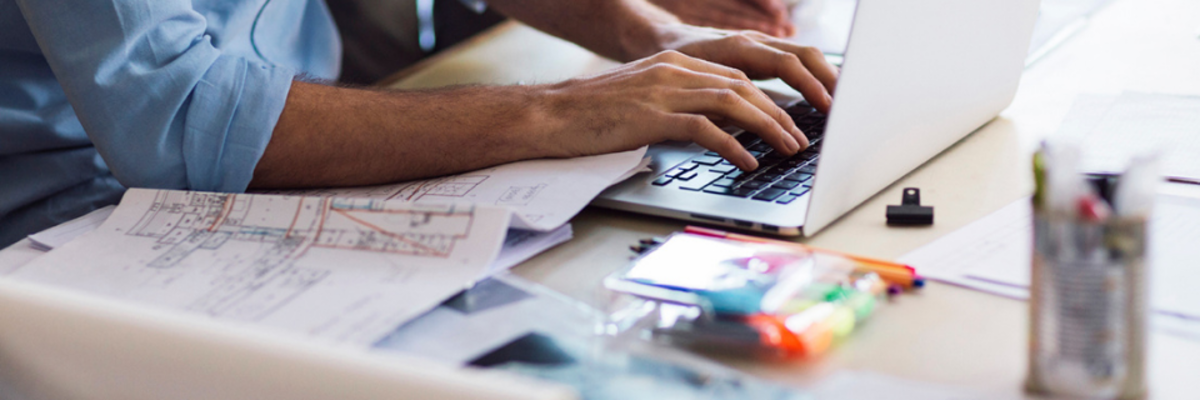 Person typing on laptop with blueprints, highlighters and design materials spread across a workspace.