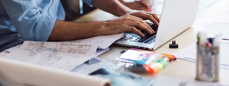 Person typing on laptop with blueprints, highlighters and design materials spread across a workspace.