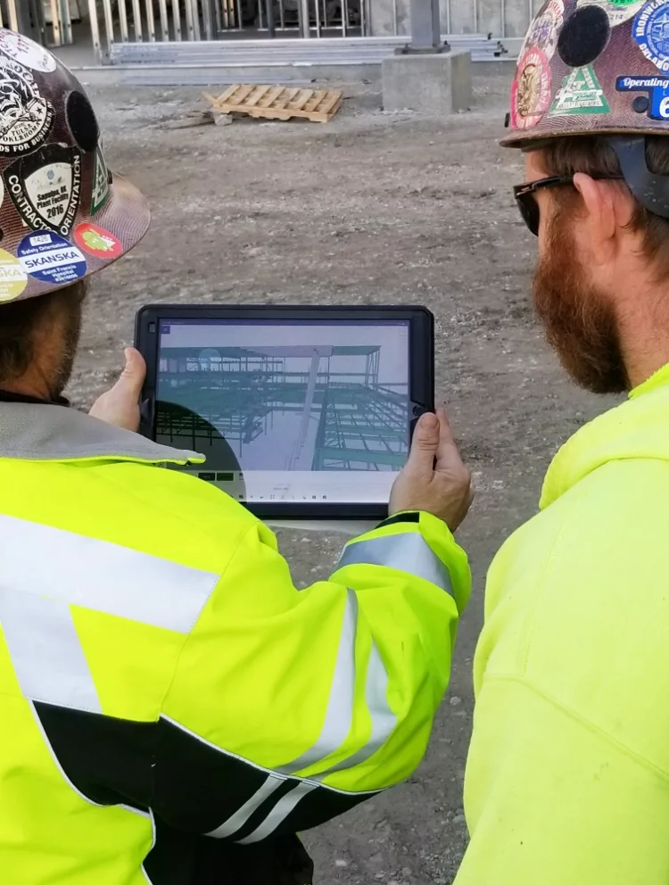 Two construction workers in hard hats and high-visibility vests look at a tablet displaying a 3D model of a building.