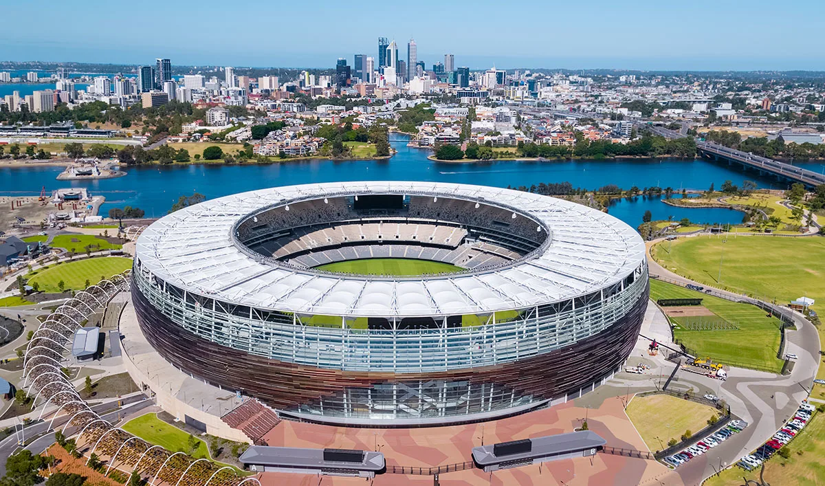An aerial view of Optus Stadium in Perth, Australia, with the city skyline and Swan River in the background.