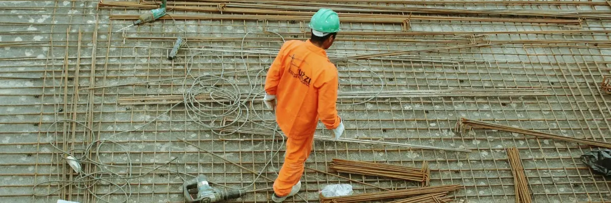 Worker in orange uniform and helmet walking on rebar grid at a construction site.