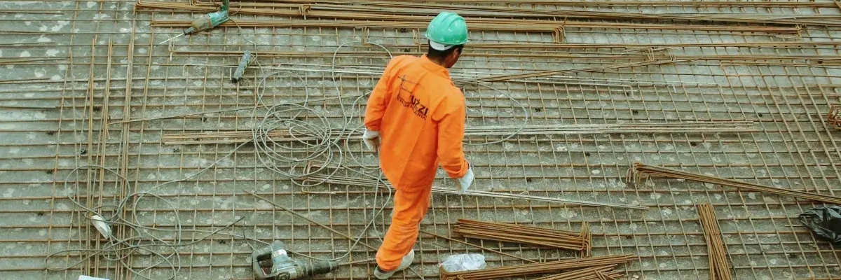 Worker in orange uniform and helmet walking on rebar grid at a construction site.