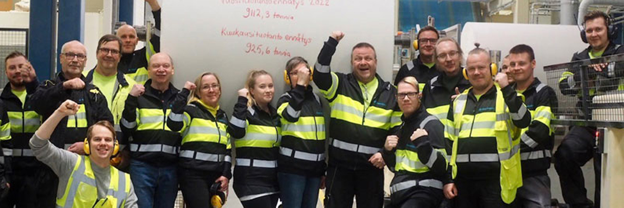 A large group of industrial workers in safety gear celebrate in front of a giant paper roll with Finnish text.