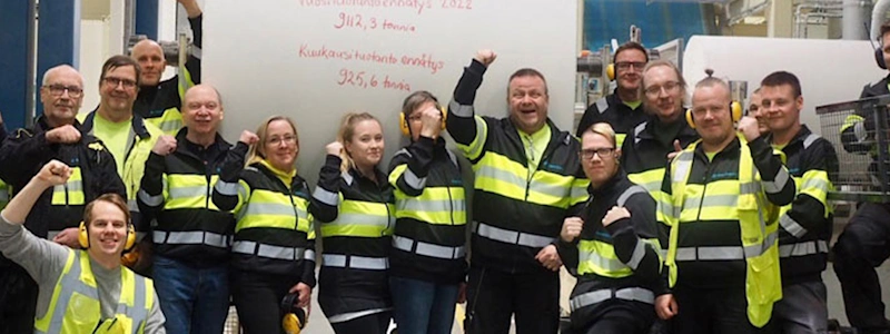 A large group of industrial workers in safety gear celebrate in front of a giant paper roll with Finnish text.