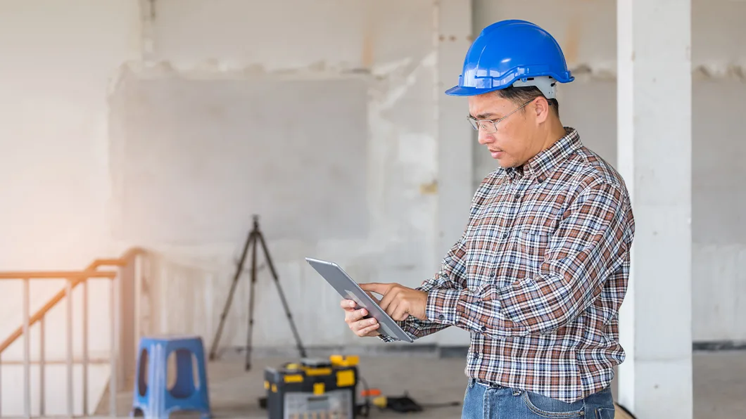 A construction worker in a blue hard hat and plaid shirt reviews a tablet at an unfinished building site.