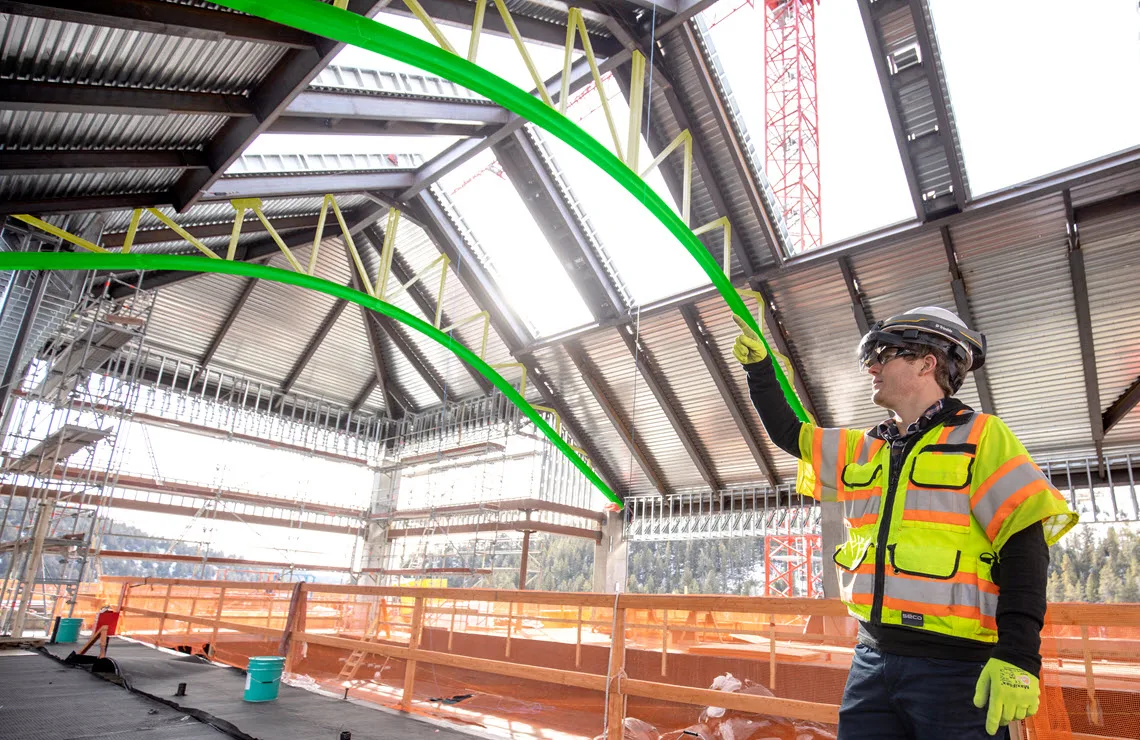 A construction worker in a bright yellow safety vest points at a large green pipe that curves across the ceiling of an unfinished building.