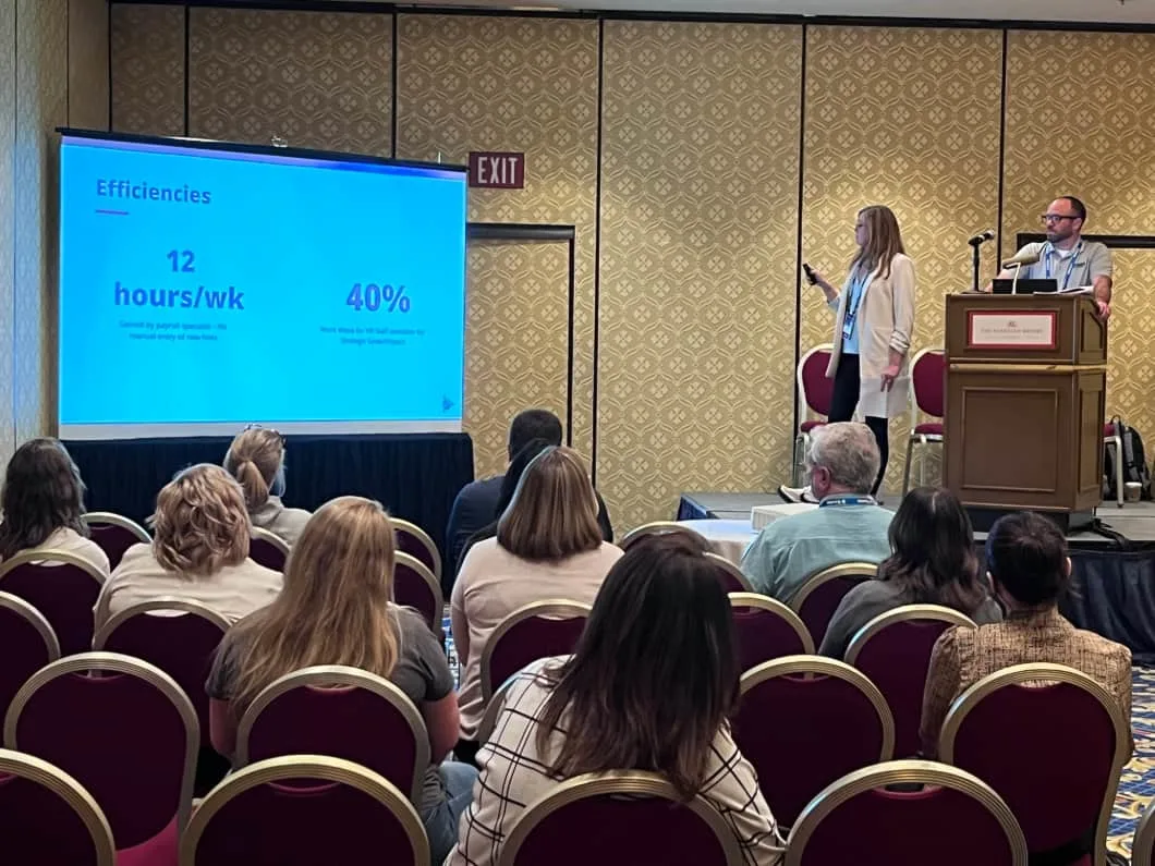 A presenter stands at a podium next to a screen displaying 12 hours/wk and 40% with an audience seated in front of them.