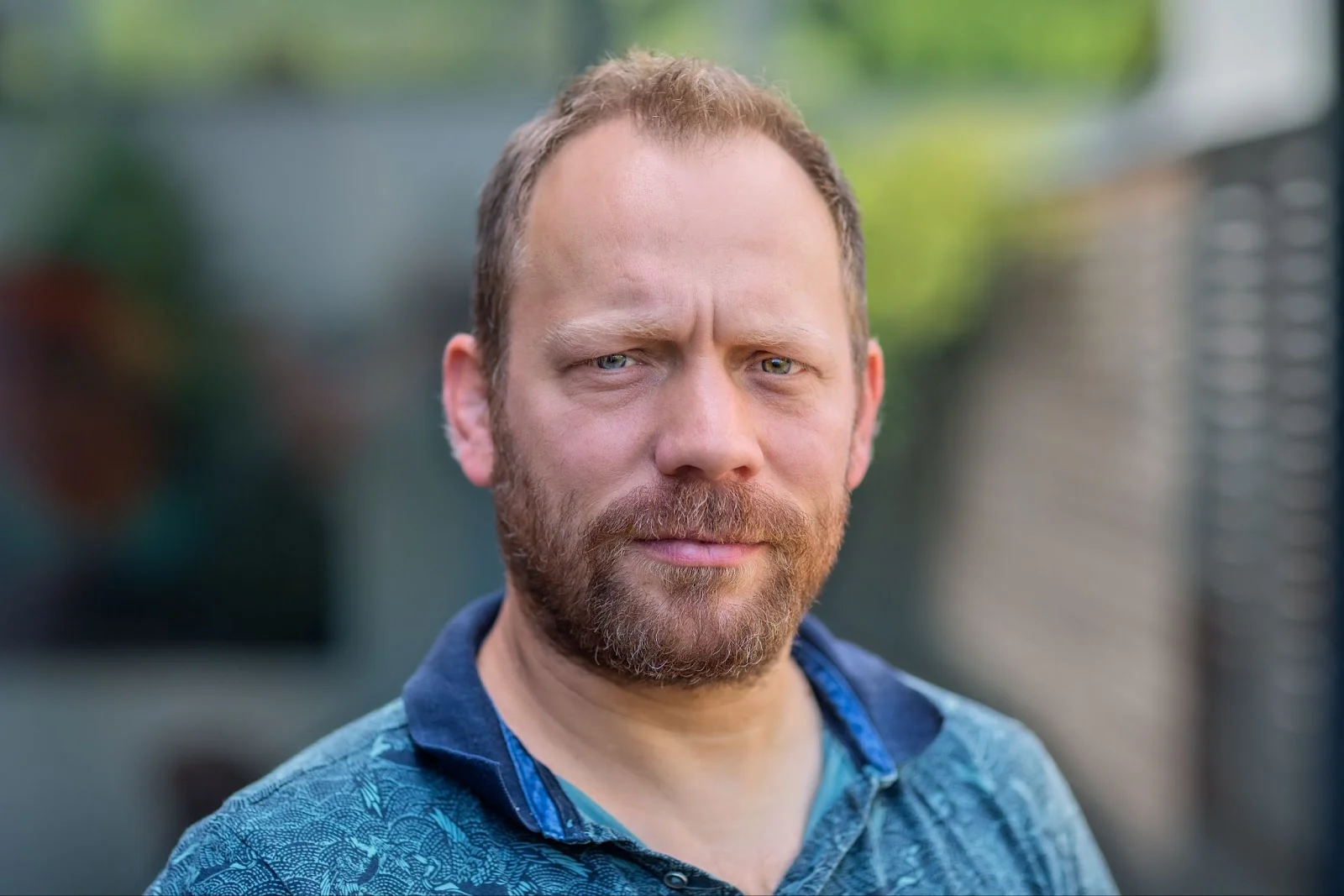Close-up portrait of a man with fair skin, light brown hair, and a beard. He is wearing a blue patterned shirt with a dark blue collar.