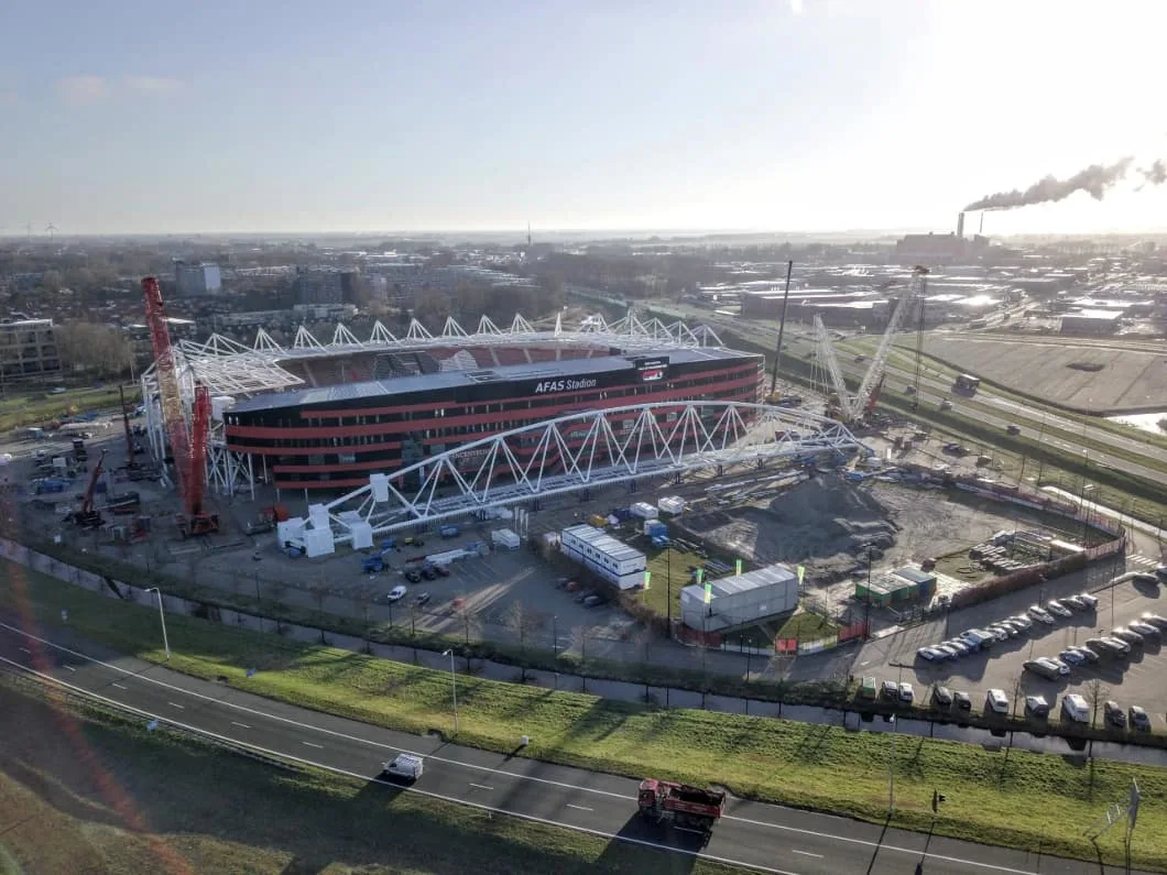 Construction of a modern stadium with red and black exterior, featuring large white truss structures and cranes on a sunny day.