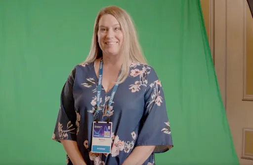 A smiling woman in a blue floral dress and conference badge stands in front of a green screen.