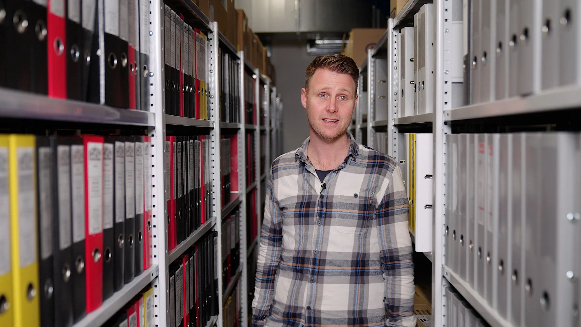 A man in a plaid shirt stands in an aisle of a large archive room, with shelves packed with binders on either side.