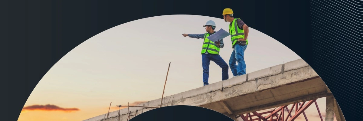 Two construction workers in yellow vests and hard hats review blueprints atop a concrete structure at sunset.