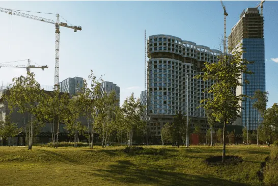 A sunny day shows construction cranes looming over a new development of curved and high-rise buildings, with a park in the foreground.