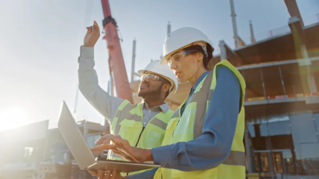Two people wearing hardhats and safety vests stand near a building construction site and note the progress of the project on their laptop.