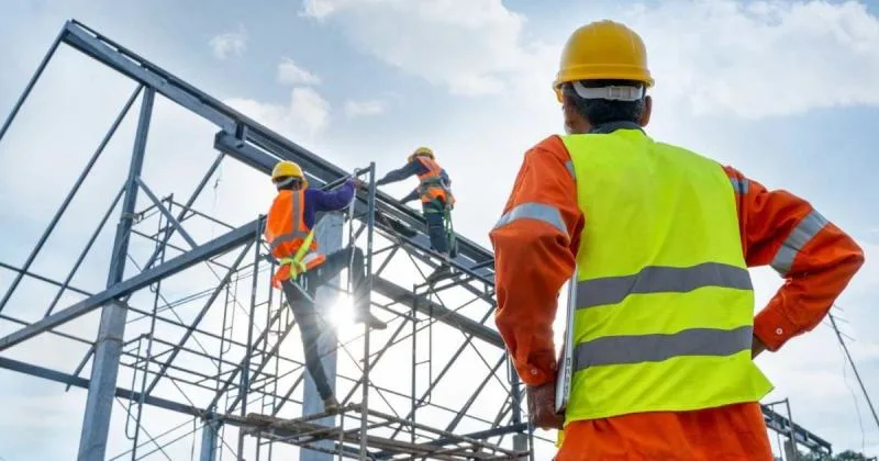 Construction workers in hard hats and high-visibility vests work on a steel frame building against a partly cloudy sky.