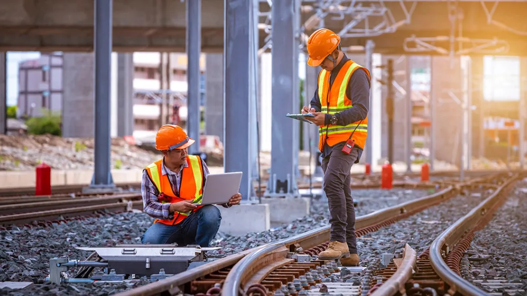 Two people wearing hardhats and safety vests at a newly constructed railway inspect the work and record measurements on their laptop and tablet.