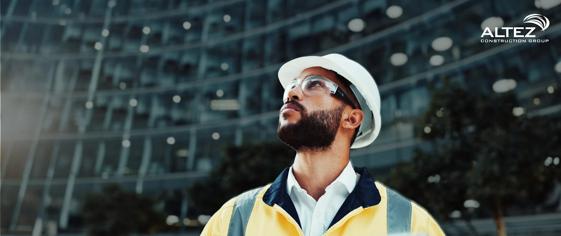 A construction worker in a yellow vest and white hard hat looks up at a large glass building while holding a tablet.