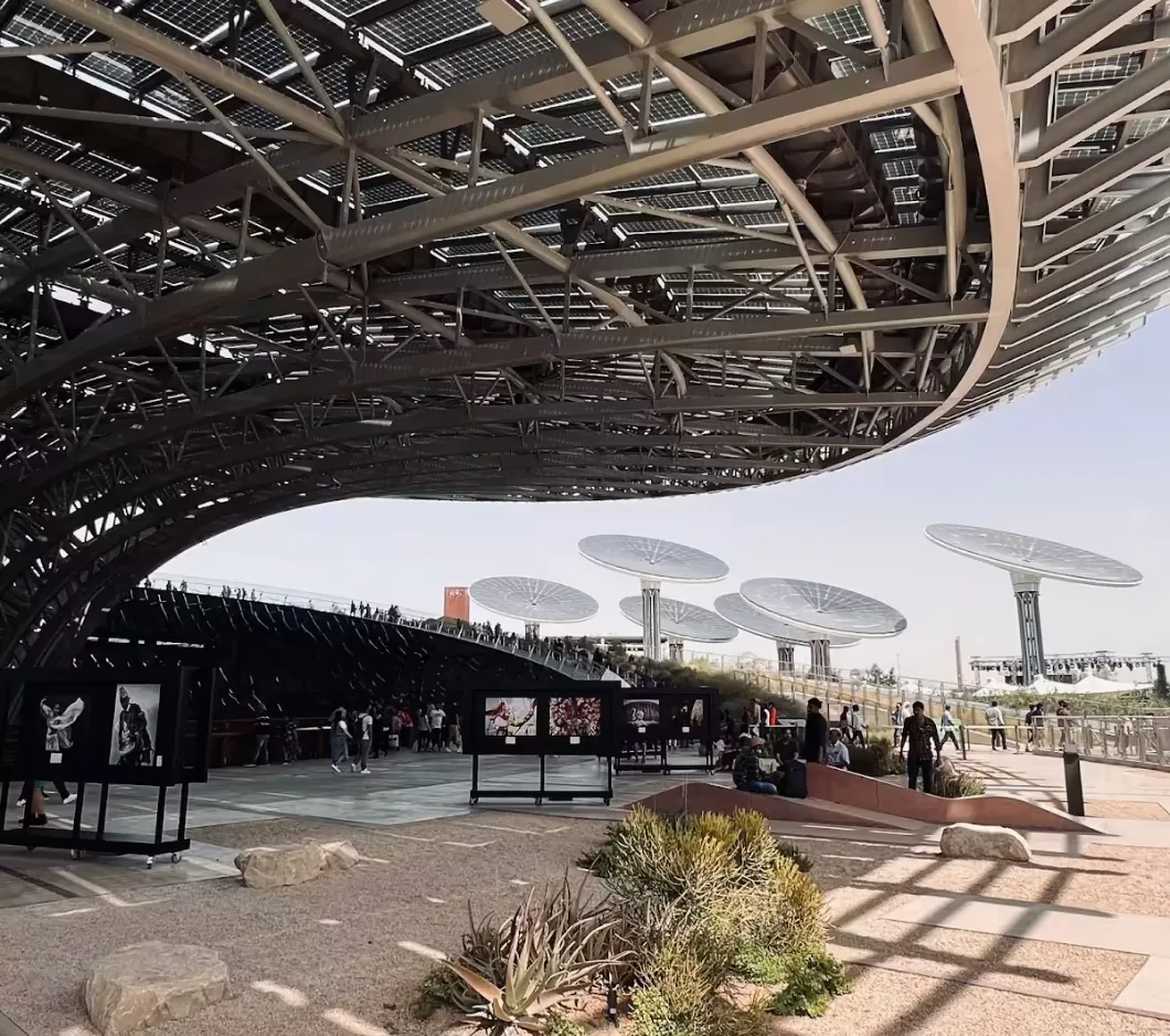 Large solar panel roof over an outdoor exhibition with people and desert plants.