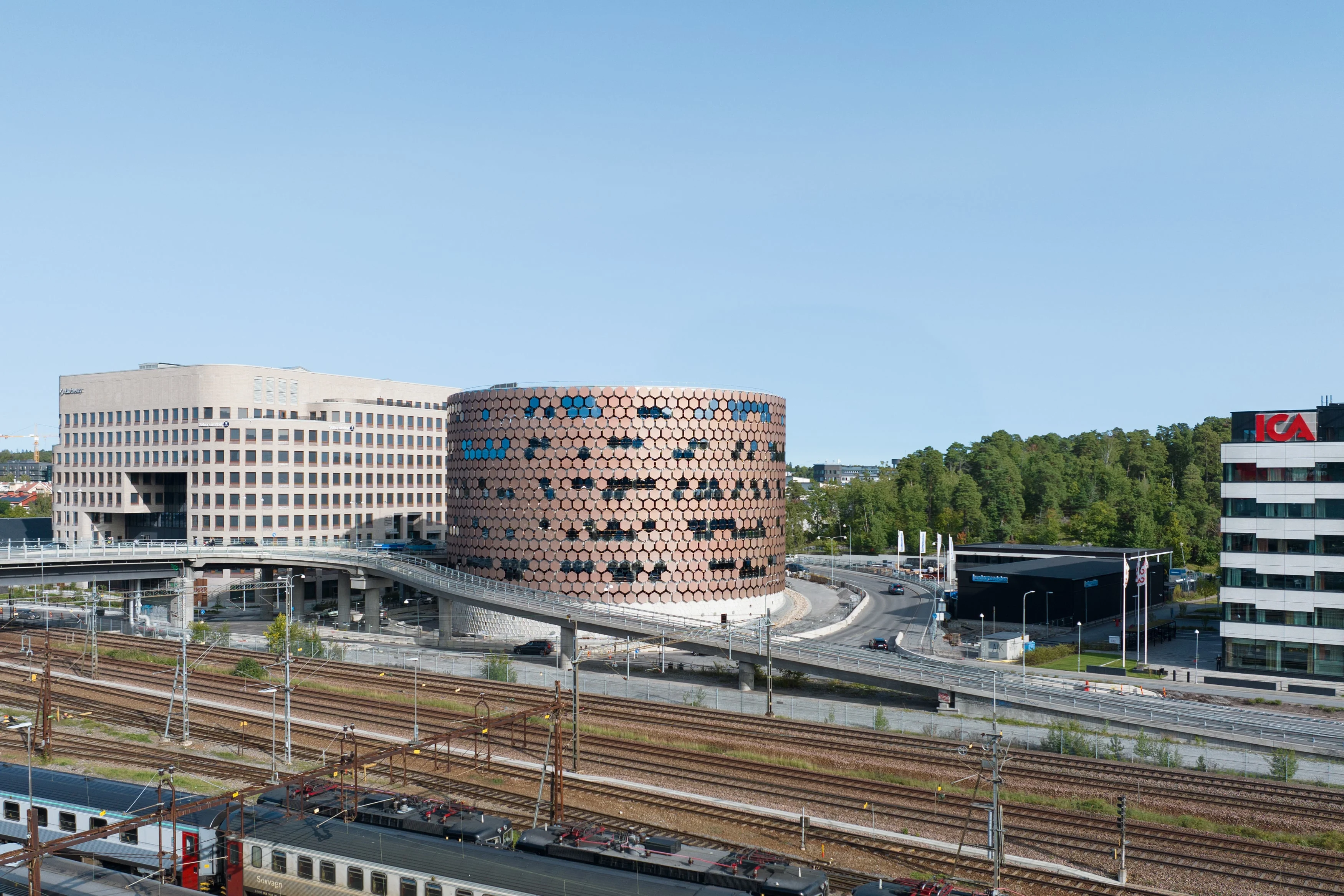 A modern cityscape, featuring a circular building with a honeycomb-like facade, adjacent to a railway line with trains.