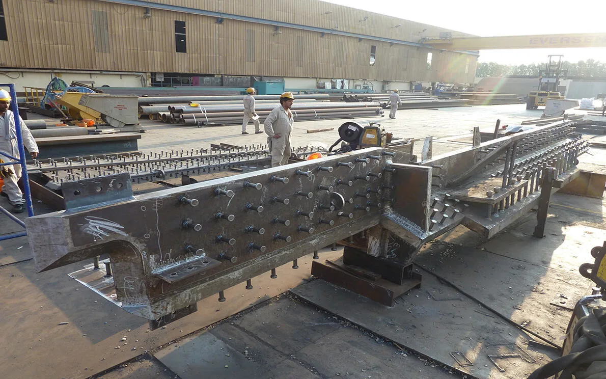 A large, custom-fabricated steel beam with numerous bolts is shown at an industrial fabrication shop.