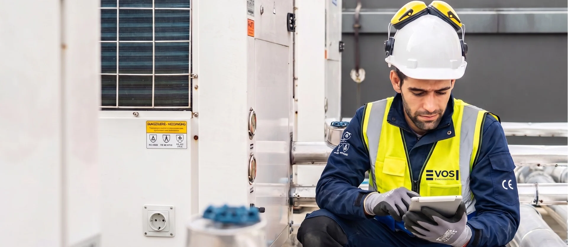 A VOS Elektrotechniek technician in a white hard hat and yellow safety vest uses a tablet near industrial HVAC equipment.