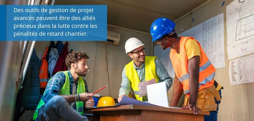 Three construction workers, wearing hard hats and safety vests, collaborate around a table with blueprints and a laptop.