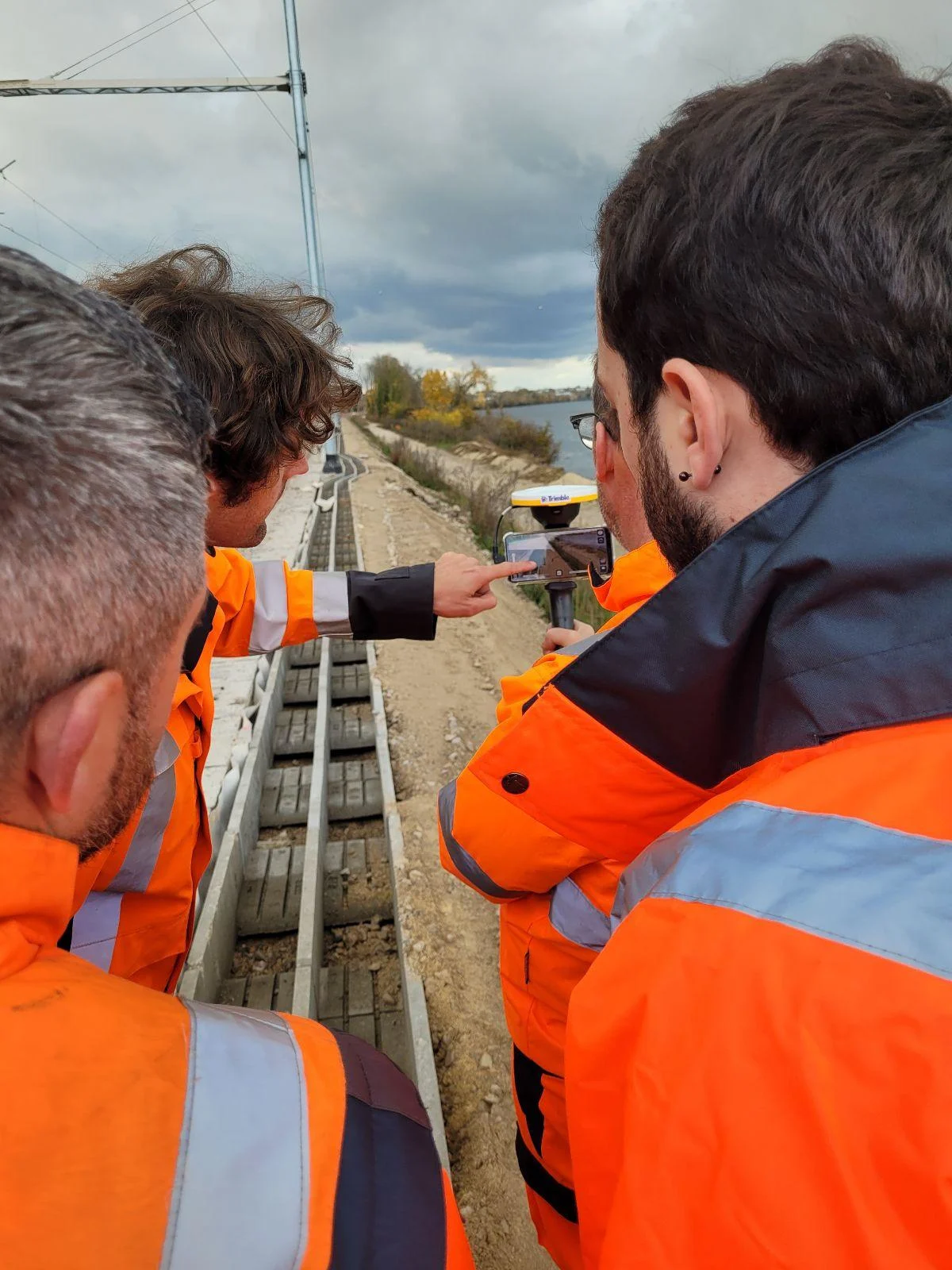 Three construction workers in orange high-visibility vests gather around a Trimble surveying device near a railway track.