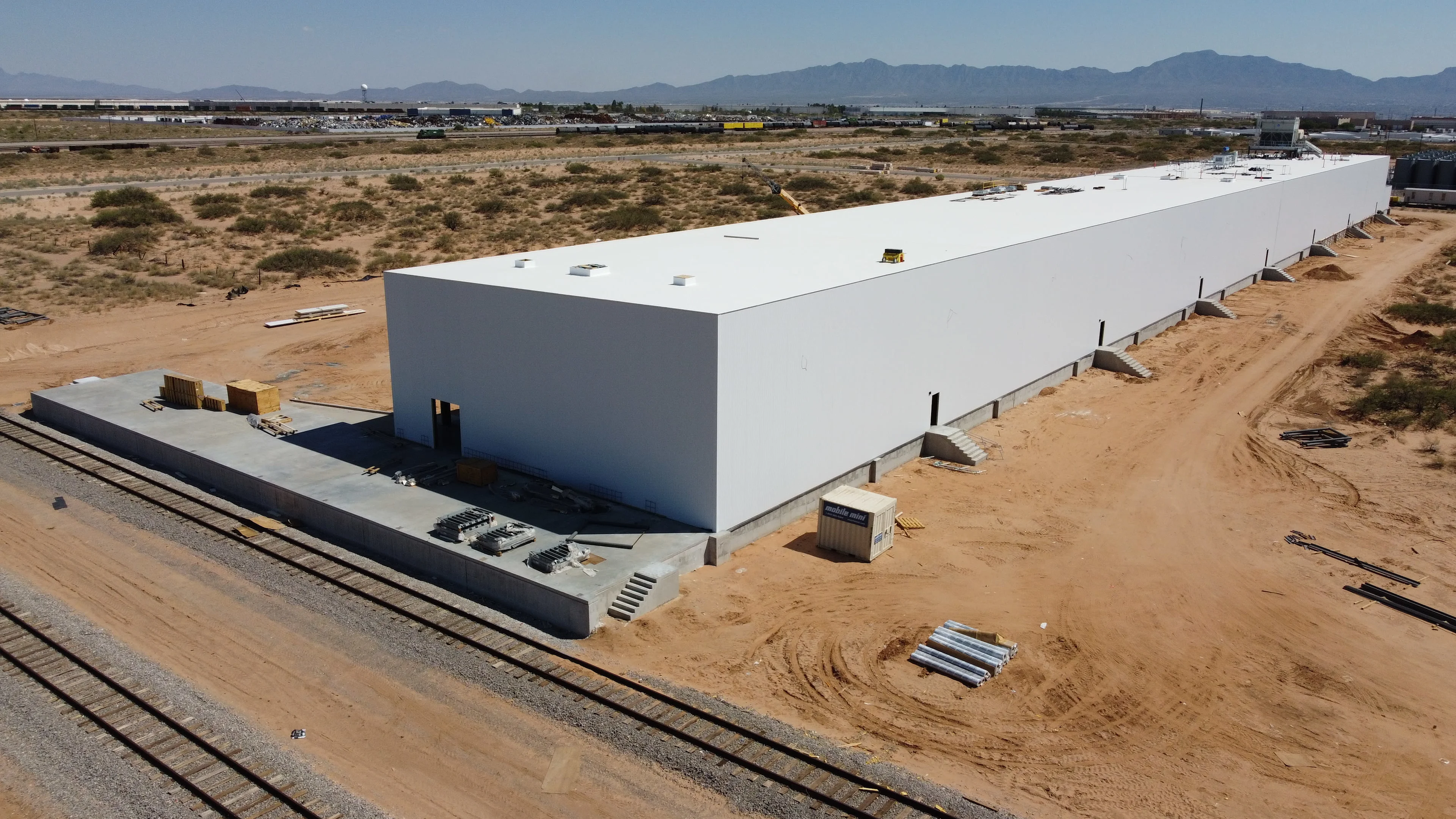 An aerial view of a long, white, modern warehouse building under construction, with railroad tracks.