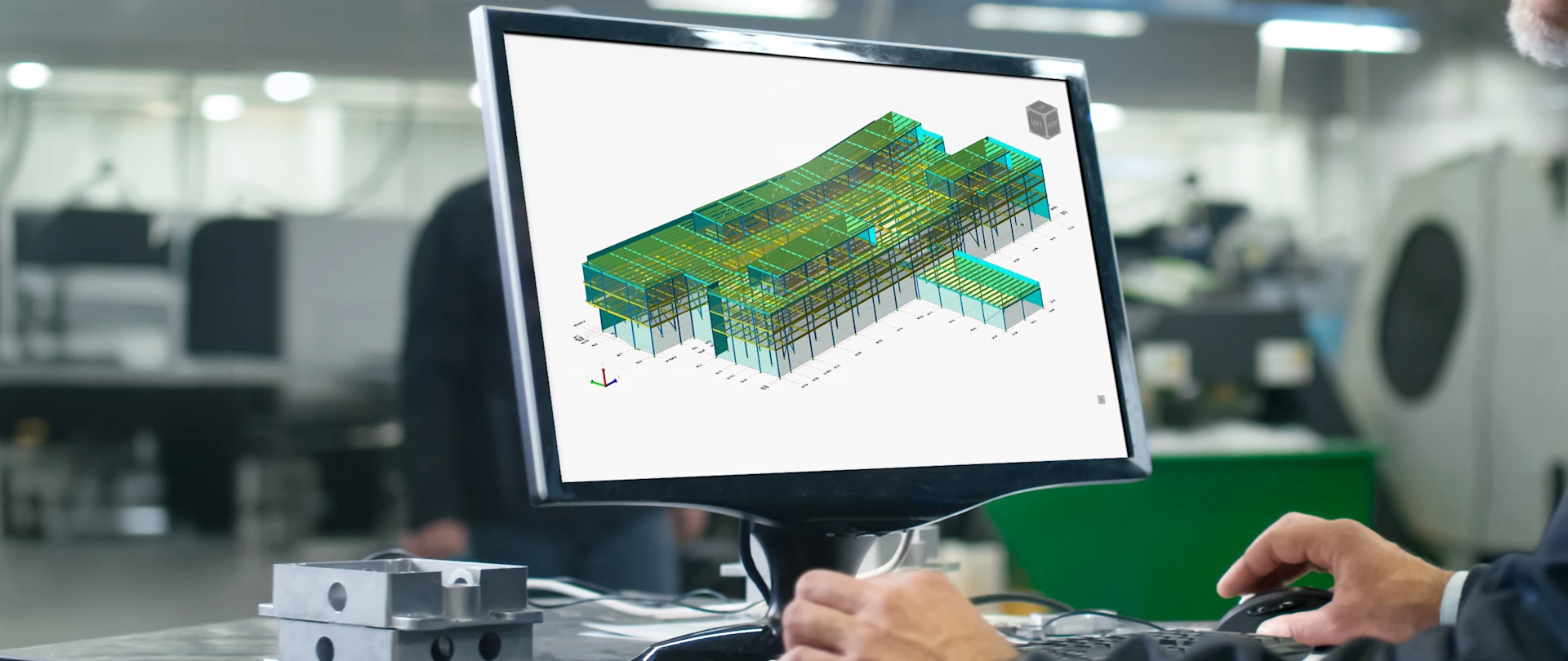 An engineer operates a computer in a modern workspace, displaying a detailed, color-coded 3D structural BIM model of a large commercial building on a desktop monitor.