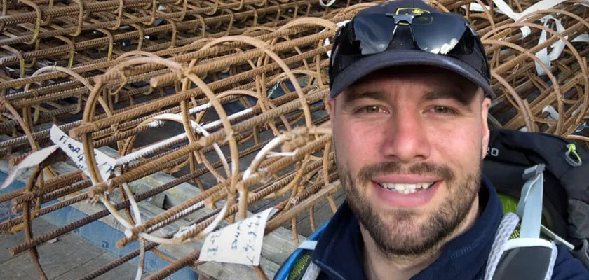 A man in a baseball cap and sunglasses smiles for a selfie in front of a pile of rebar.