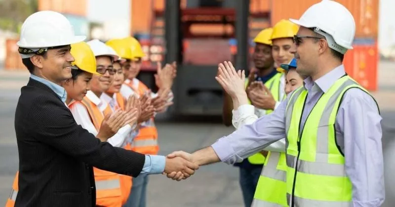 Two men in hard hats shake hands, while a diverse group of workers in safety vests and hard hats applaud behind them.