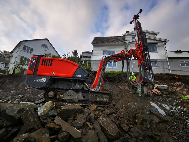 A construction worker operates a drilling machine near houses on a rocky slope.