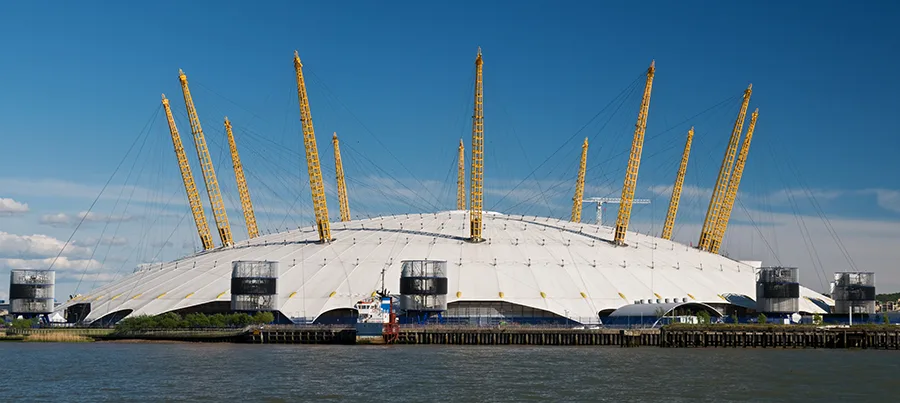 The O2 Arena, a large domed entertainment venue, stands by the river under a clear blue sky.