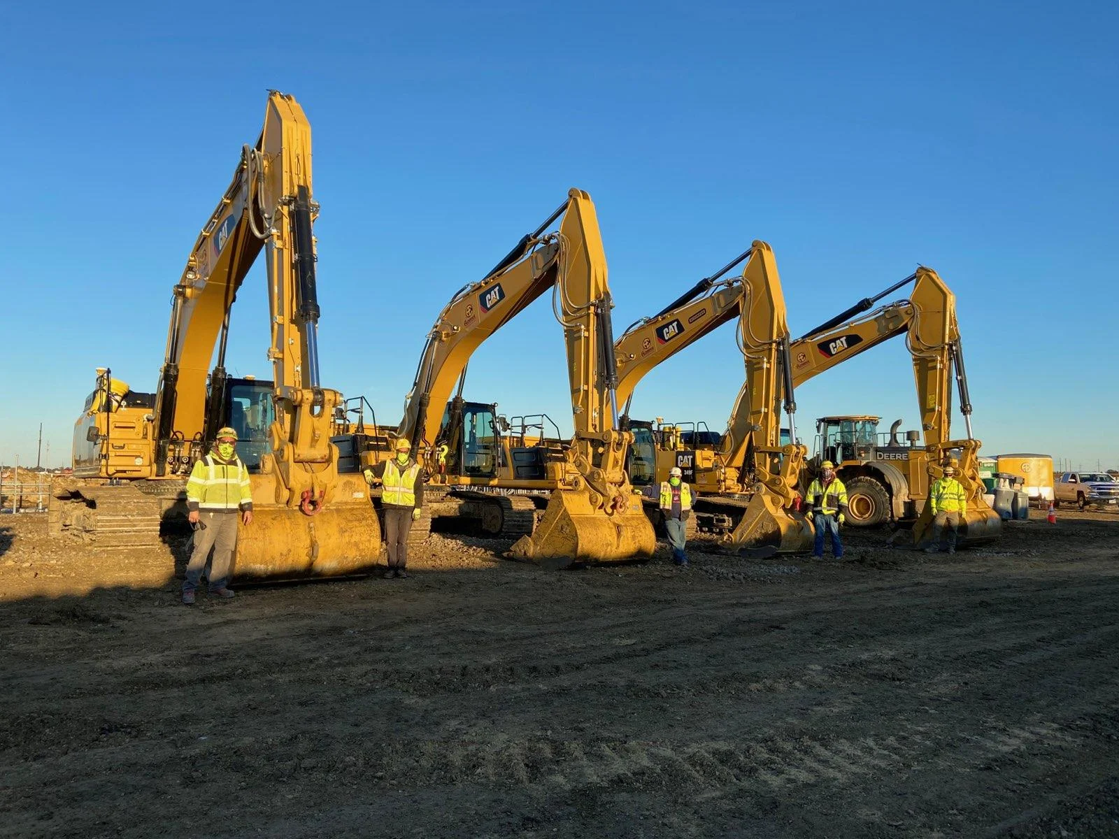 A group of workers stand in front of several yellow excavators and a loader under a clear blue sky.