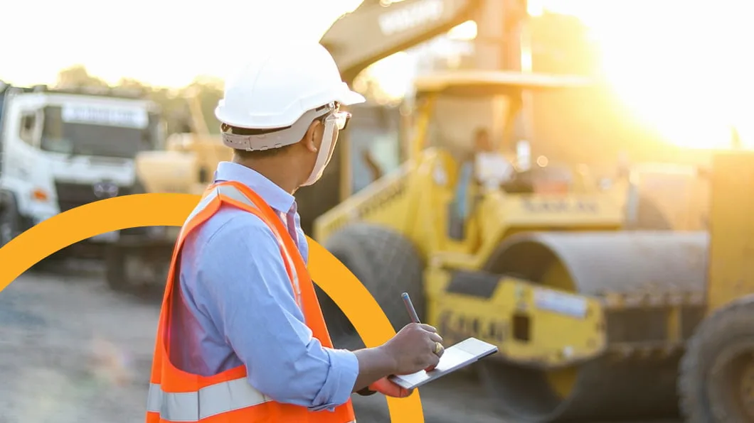 An engineer in a hard hat and safety vest uses a tablet while inspecting a concrete bridge construction site.