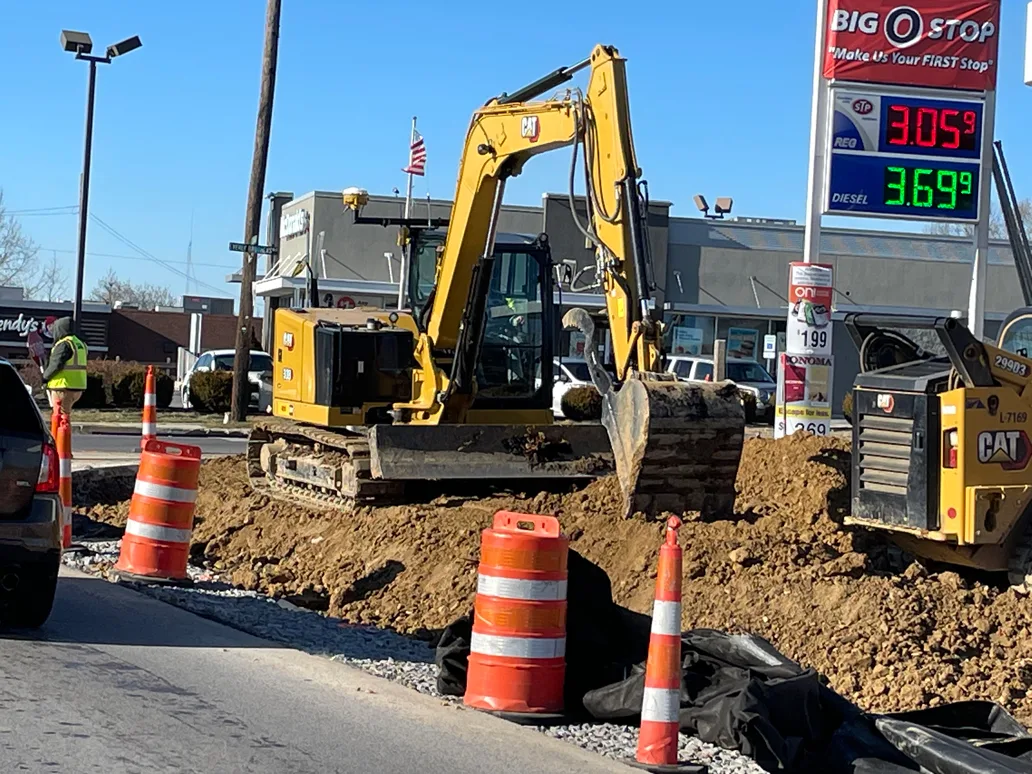 Excavator digging at a construction site next to a Big Stop gas station with gas prices displayed.
