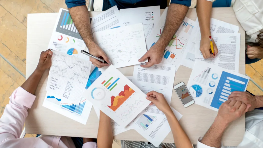 A diverse group of people collaborate around a table, analyzing charts and data.