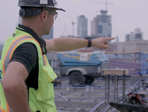 Construction worker in hard hat and vest points with his finger toward the city skyline.