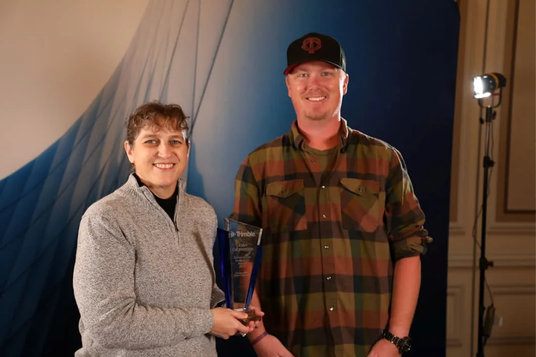 A woman in a grey quarter-zip sweater and a man in a plaid shirt and baseball cap smile as they pose with a blue glass award.