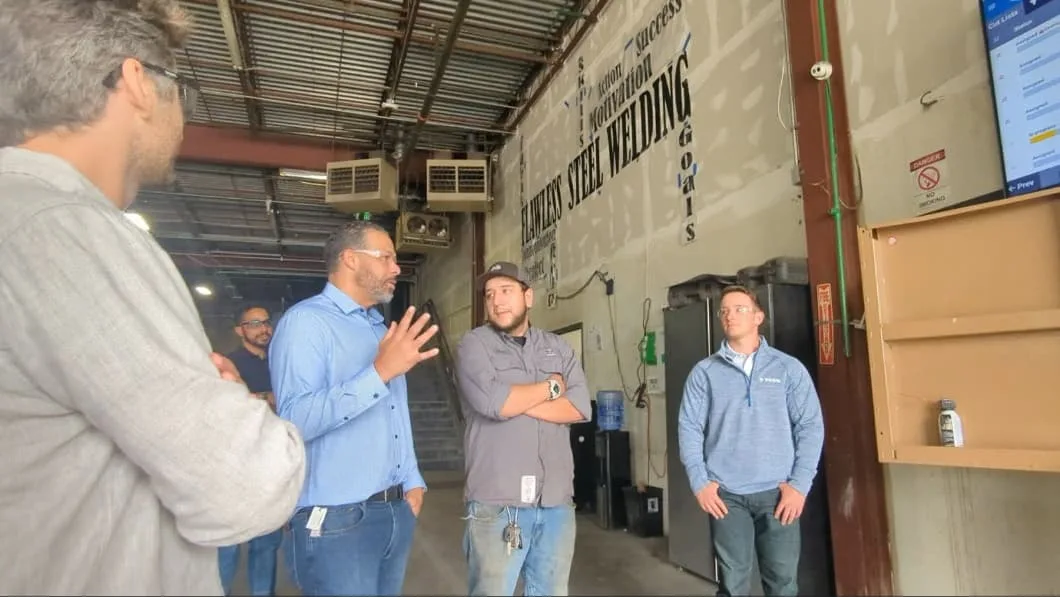 A group of men stand in a workshop, listening to a speaker. Motivational words are painted on the wall behind them.