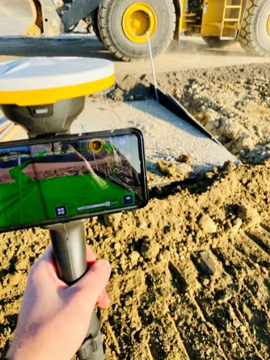 A person holds a smartphone displaying an AR overlay over a construction site.