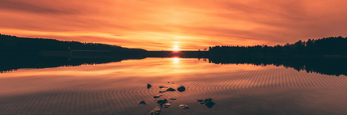 A concrete dam or water management infrastructure is silhouetted against a vibrant sunset over a lake, reflecting the orange sky on rippled water, indicating an active civil engineering project.