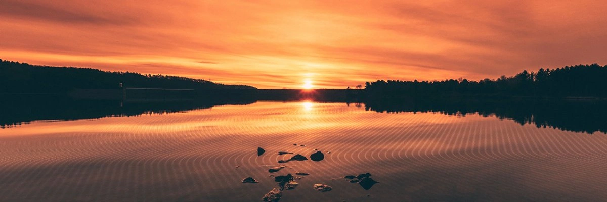 A concrete dam or water management infrastructure is silhouetted against a vibrant sunset over a lake, reflecting the orange sky on rippled water, indicating an active civil engineering project.
