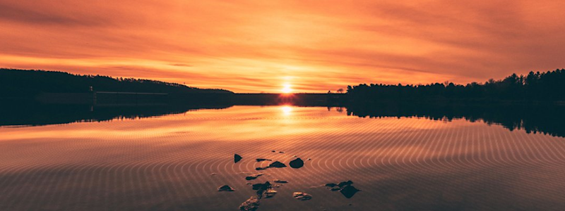 A concrete dam or water management infrastructure is silhouetted against a vibrant sunset over a lake, reflecting the orange sky on rippled water, indicating an active civil engineering project.