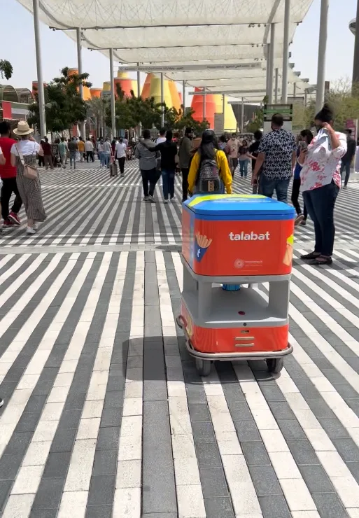 A delivery robot with talabat branding navigates a busy pedestrian walkway with striped pavement.