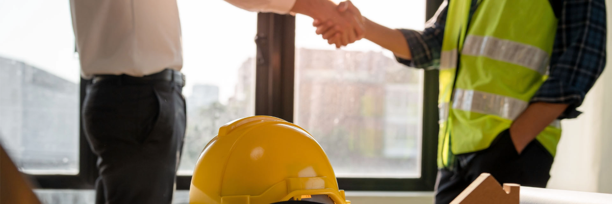 Deux personnes se serrent la main dans un bureau, avec un casque de chantier posé sur la table.