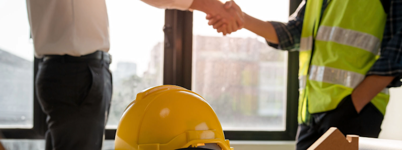 Deux personnes se serrent la main dans un bureau, avec un casque de chantier posé sur la table.